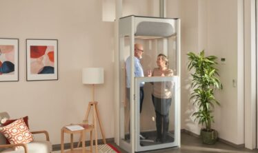 Ηλικιωμένο ζευγάρι σε έναν ανελκυστήρα σπιτιού της Stannah Uplifts σε ένα φωτεινό σαλόνι που παρουσιάζει συμπαγή κατασκευή ασανσέρ με διάφανα πάνελ.