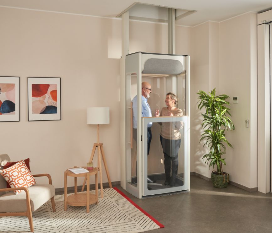 Ηλικιωμένο ζευγάρι σε έναν ανελκυστήρα σπιτιού της Stannah Uplifts σε ένα φωτεινό σαλόνι που παρουσιάζει συμπαγή κατασκευή ασανσέρ με διάφανα πάνελ.