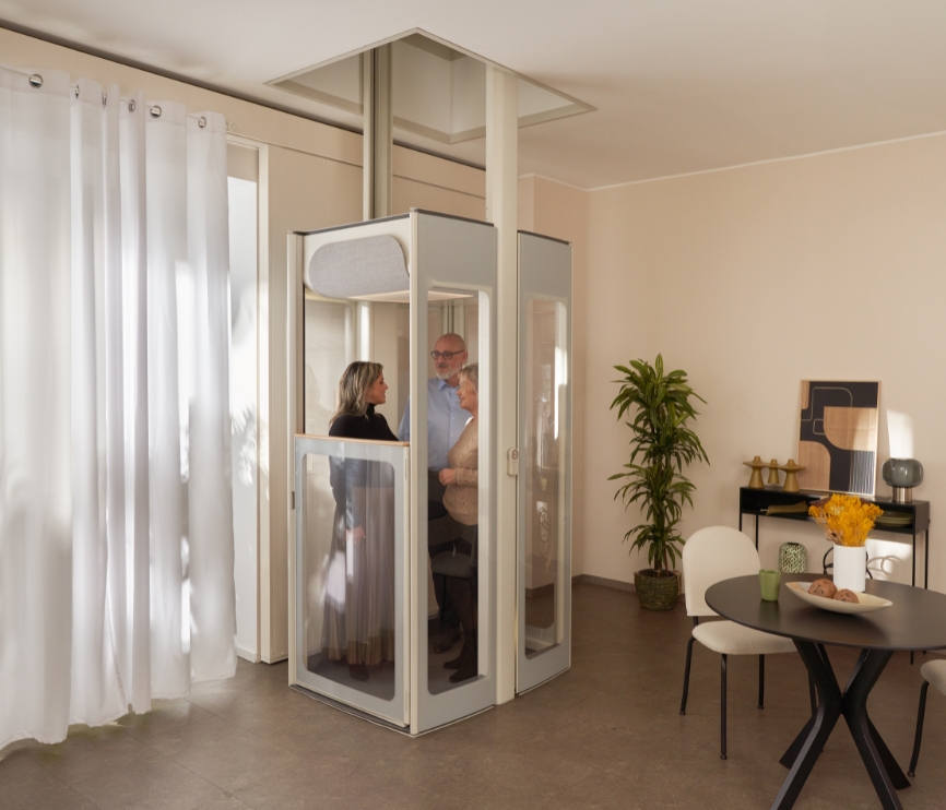 Τρεις ενήλικες στέκονται μέσα σε έναν οικιακό ανελκυστήρα με διαφανή πάνελ σε έναν μοντέρνο χώρο τραπεζαρίας και καθιστικού.